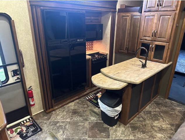 A modern RV kitchen featuring a large black refrigerator, stone-look countertops, a sink, and wooden cabinetry.