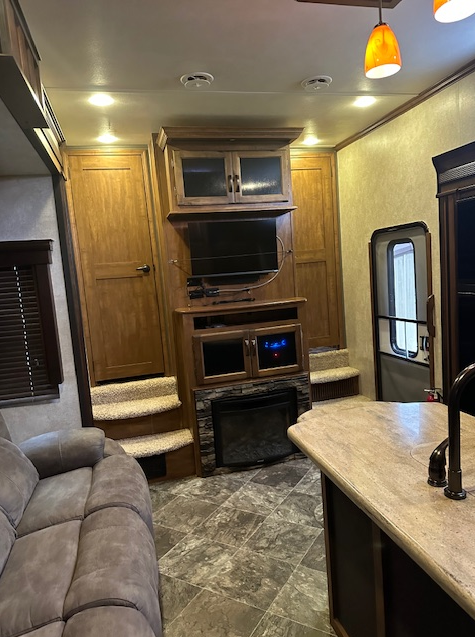 A modern RV interior featuring a gray sofa, a wood entertainment center with a TV and fireplace, and a kitchen island.