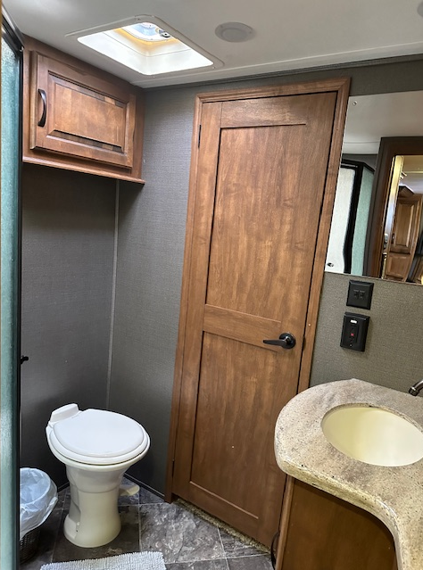 A bathroom in an RV featuring a white toilet, wood cabinetry, a sink with a beige countertop, and a skylight.