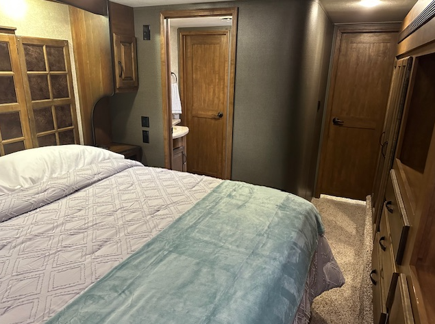 A bedroom with a bed topped with a light grey patterned comforter and a teal blanket, leading to an open doorway.