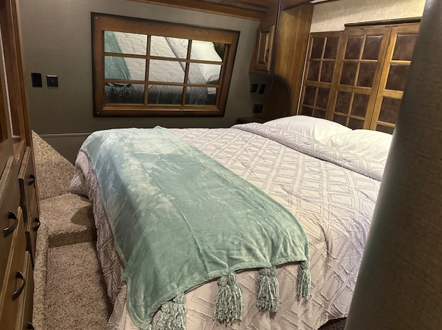 A bed with a light blue throw blanket and white quilted bedding, set in a wood-paneled room with a window.