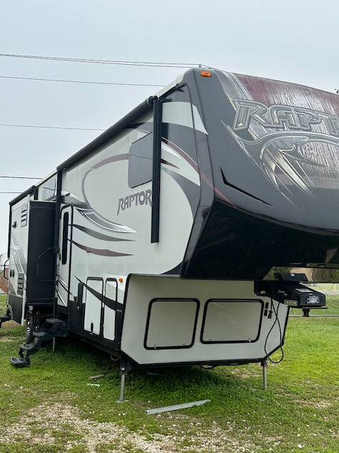 A white and black Raptor fifth-wheel RV parked on a grassy field under a cloudy sky.