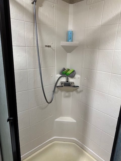 Small white corner shower with a handheld showerhead and toiletries.