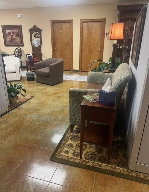 A lobby with tan tile, a gray partition, a patterned loveseat with a brown seat, a side table with a lamp, and a saddle.