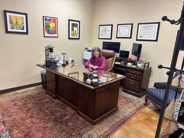 Woman in an office smiling, seated behind a large wooden desk. Art and diplomas on wall.