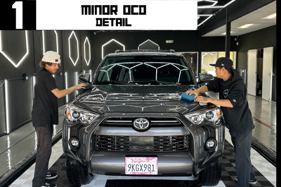 Two people wash a dark gray Toyota SUV. 