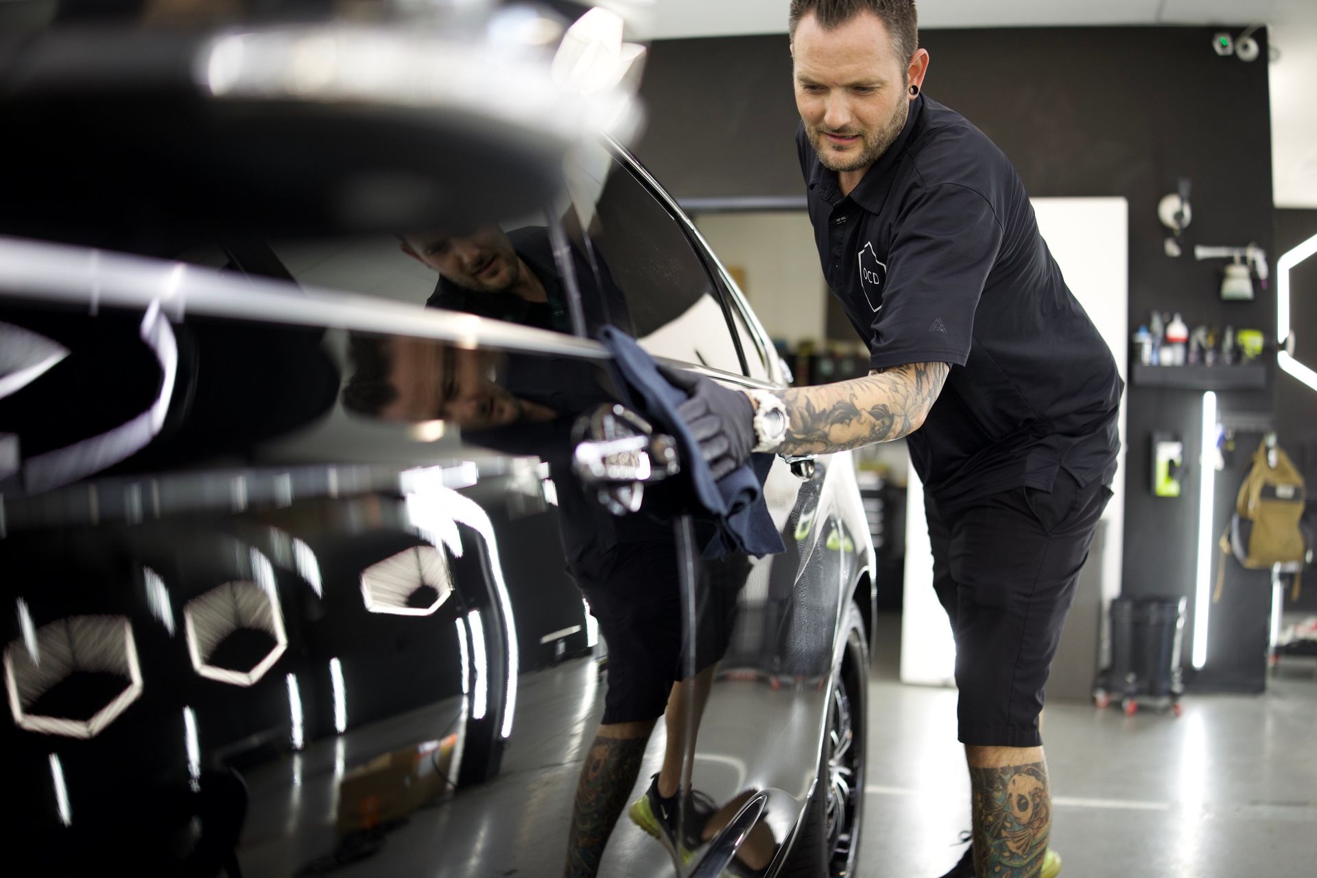 A technician wearing a black uniform and tattoos on their arms polishes the glossy black exterior of a car in a shop.