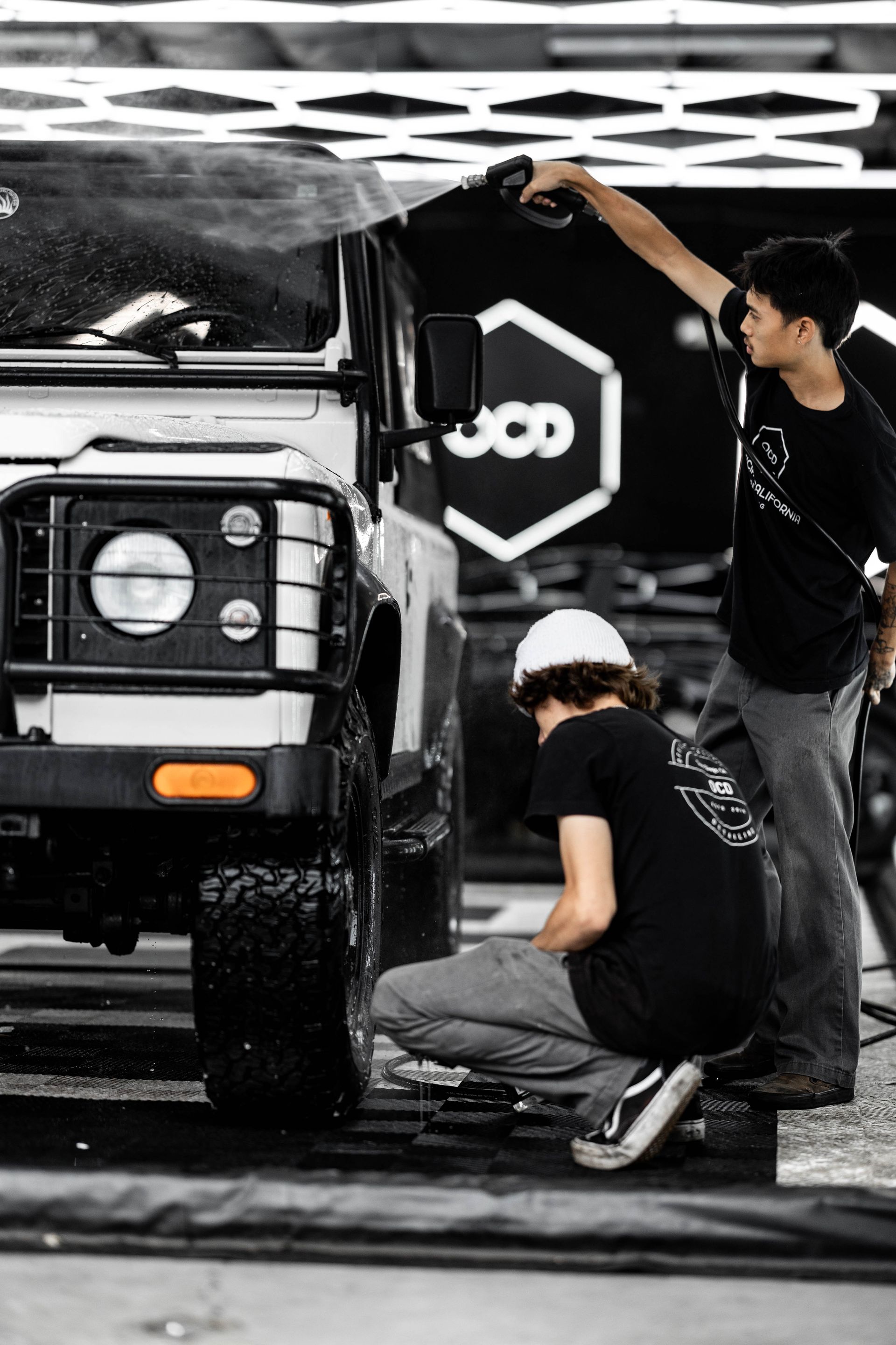 Two people washing a Land Rover. One on the tire, the other spraying the windshield, in a service bay.