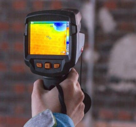 Handheld thermal camera aimed at a brick wall, showing a heat map in yellow, orange, and blue.