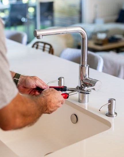 Hands using red pliers to tighten a chrome faucet on a white bathroom sink.