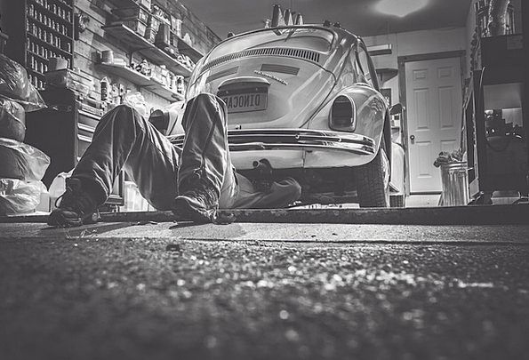 Person lying under a classic car in a cluttered garage, viewed from ground level.