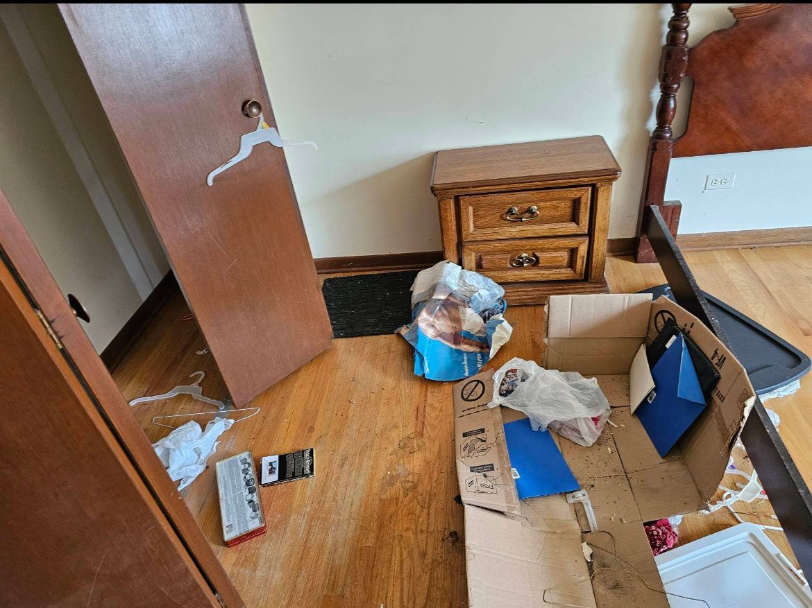 Messy room with an open door, scattered boxes, papers, and a blue bag on the floor beside a wooden nightstand