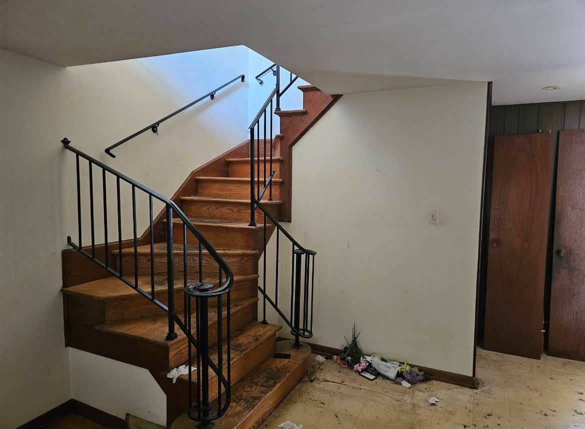 Wooden staircase with black railing in a plain indoor hallway, next to a closed door and debris on the floor