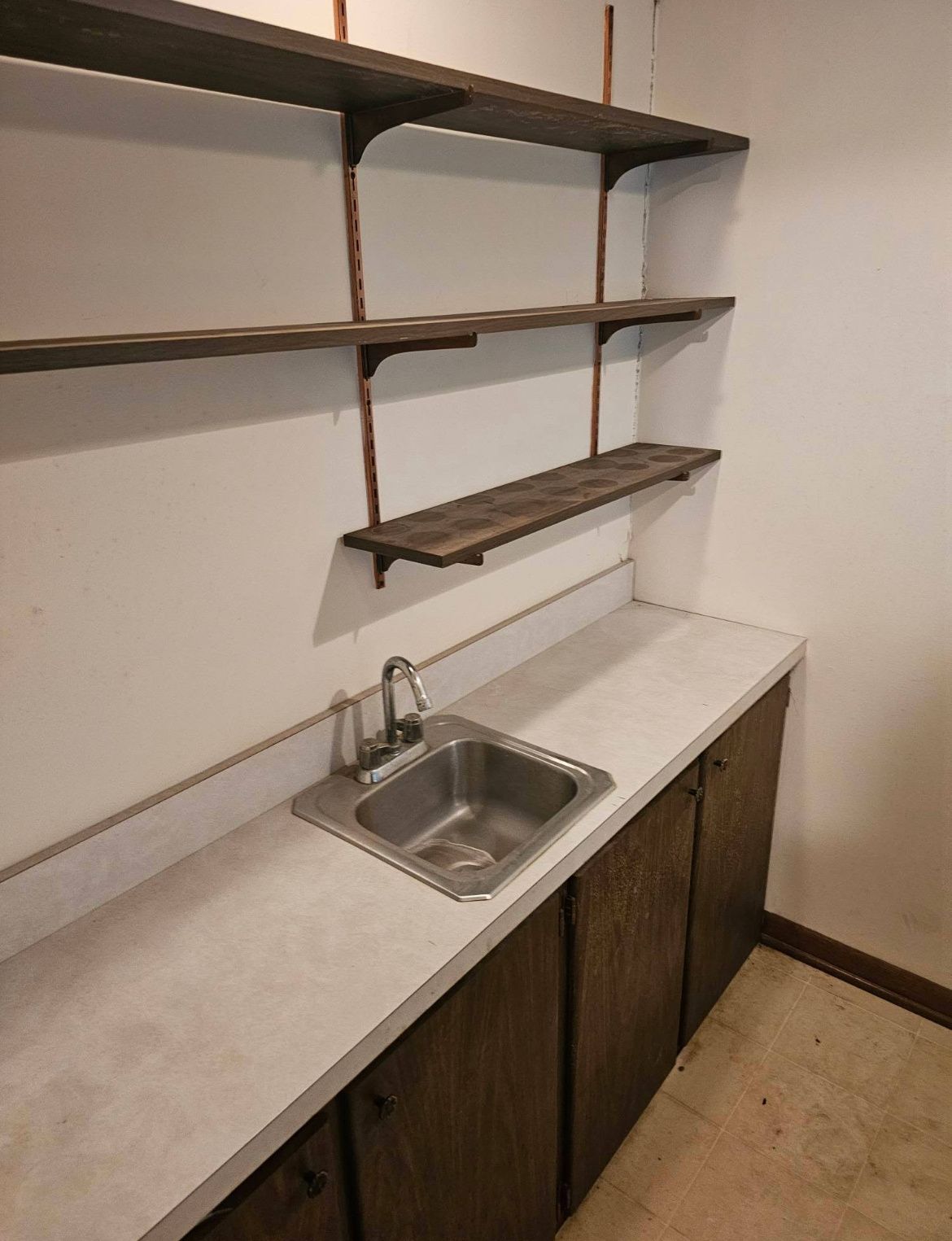Small kitchen sink and countertop with open wooden shelves on a bare wall
