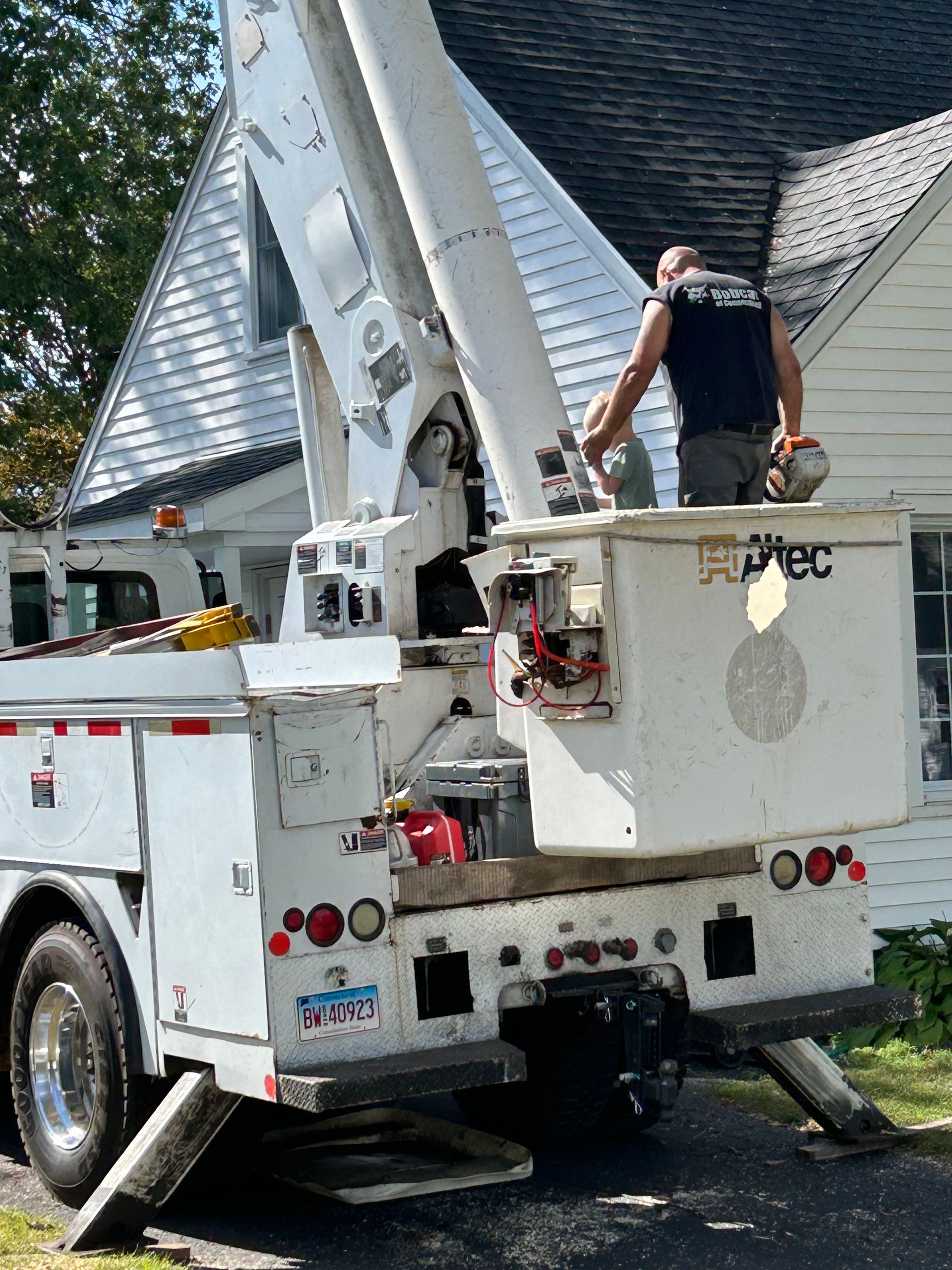 White utility bucket truck with raised boom parked by a house in a driveway, worker in the bucket.