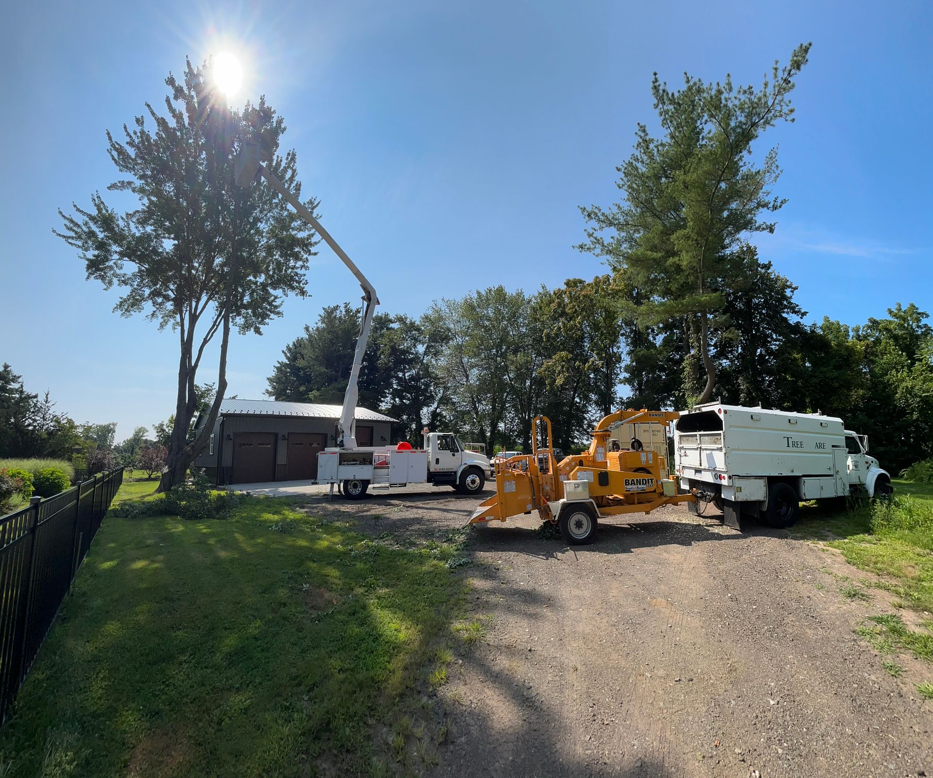 Utility trucks and equipment on a gravel driveway under bright sun, with trees and a house nearby