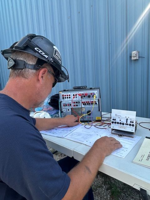 Man in a hard hat working on electrical equipment and blueprints at a table outdoors.