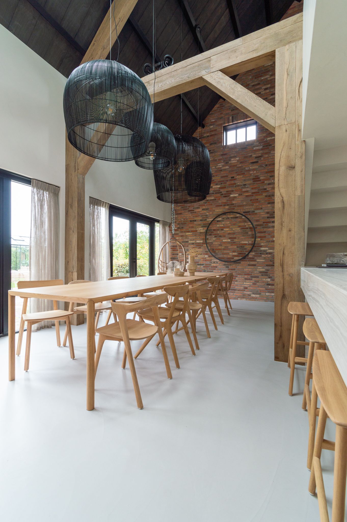Een eetkamer met een lange tafel, stoelen van Ethnicraft en een bakstenen muur. Foto uit The Art of Living.
