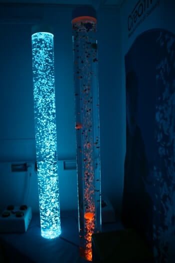 Two illuminated bubble tubes stand vertically in a dark room; one glows bright blue and the other contains red-tinted light.
