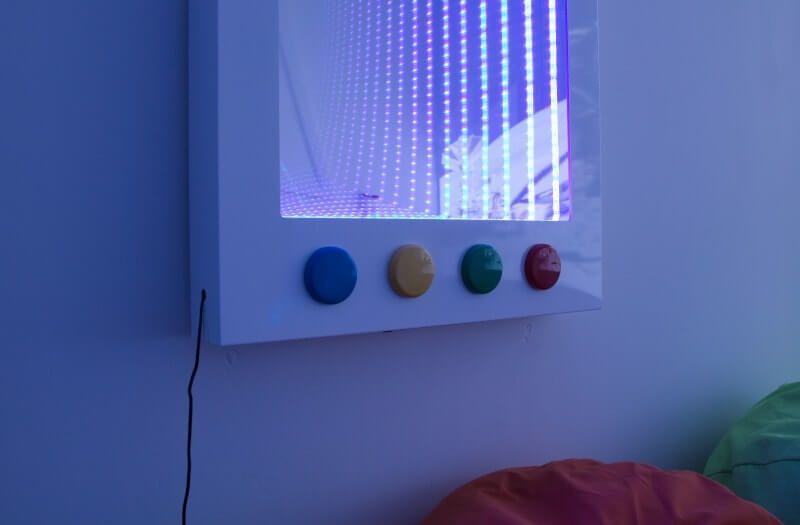 A wall-mounted infinity mirror with four colored buttons—blue, yellow, green, and red—positioned below it.
