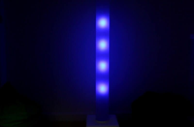 A tall, vertical tower lamp glows with four bright blue lights against a dark wall in a dimly lit room.