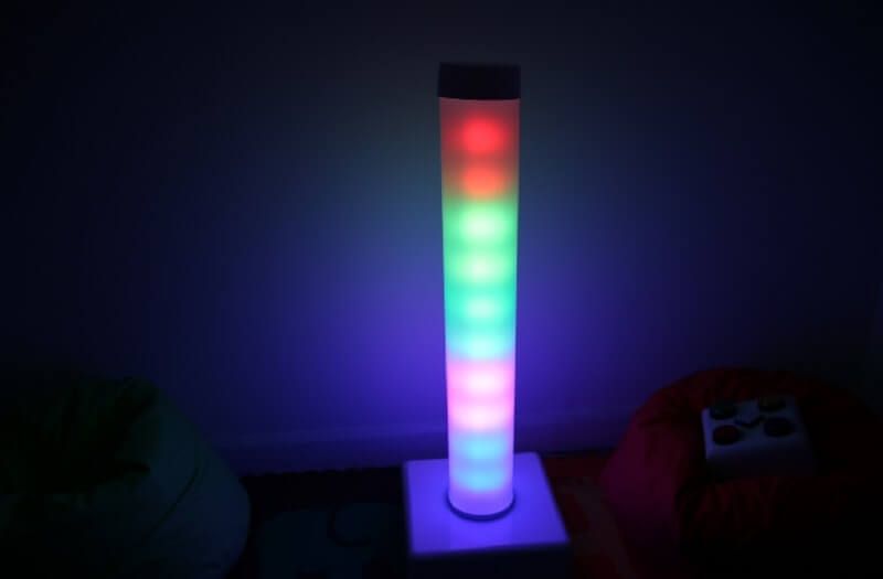 A tall, cylindrical lamp glowing in a gradient of red, green, and blue sits on a square base in a dark room.