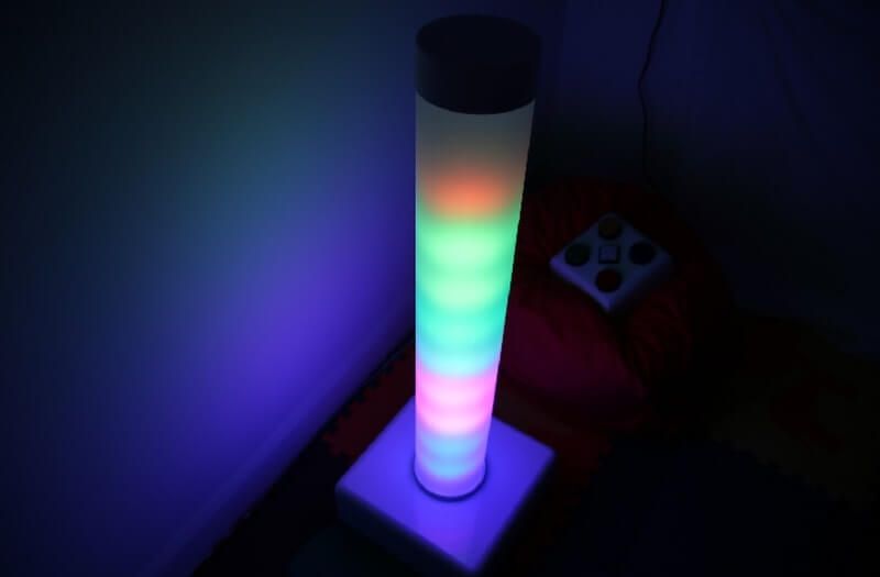 A tall, cylindrical lamp glowing in a gradient of red, green, and blue sits on a square base in a dark room.
