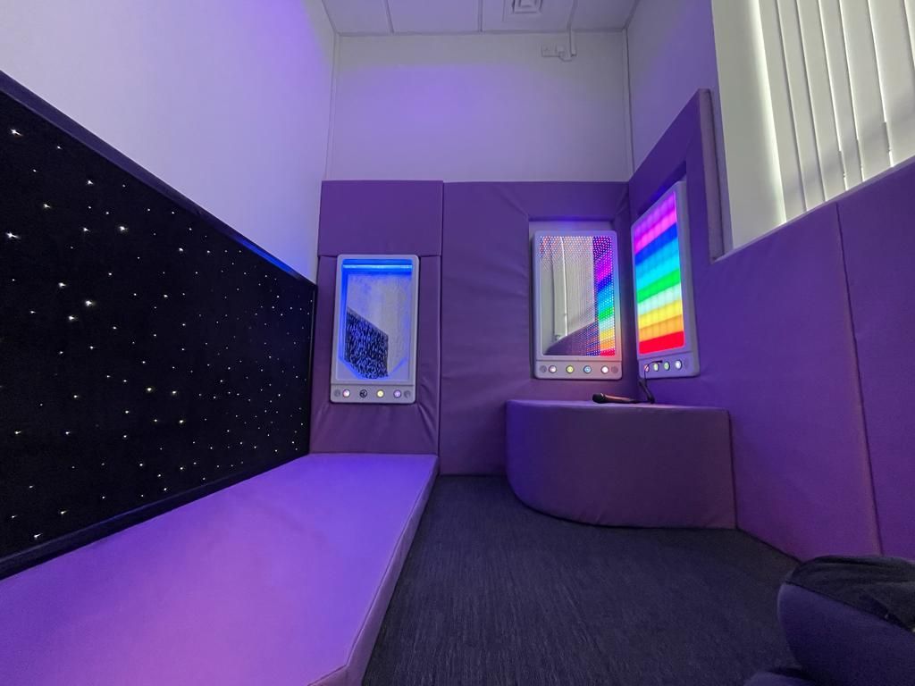 A purple sensory room featuring a starry wall panel, illuminated displays, padded seating, and a darkened, calm atmosphere.
