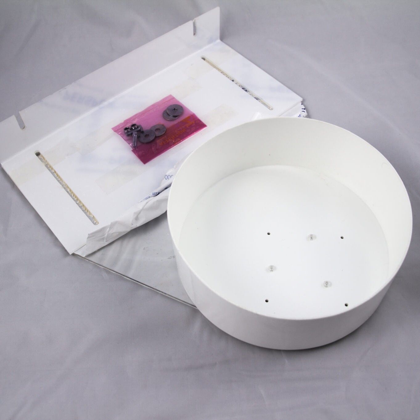 A white, round light fixture base sits next to a flat mounting bracket and a small bag of screws on a gray surface.