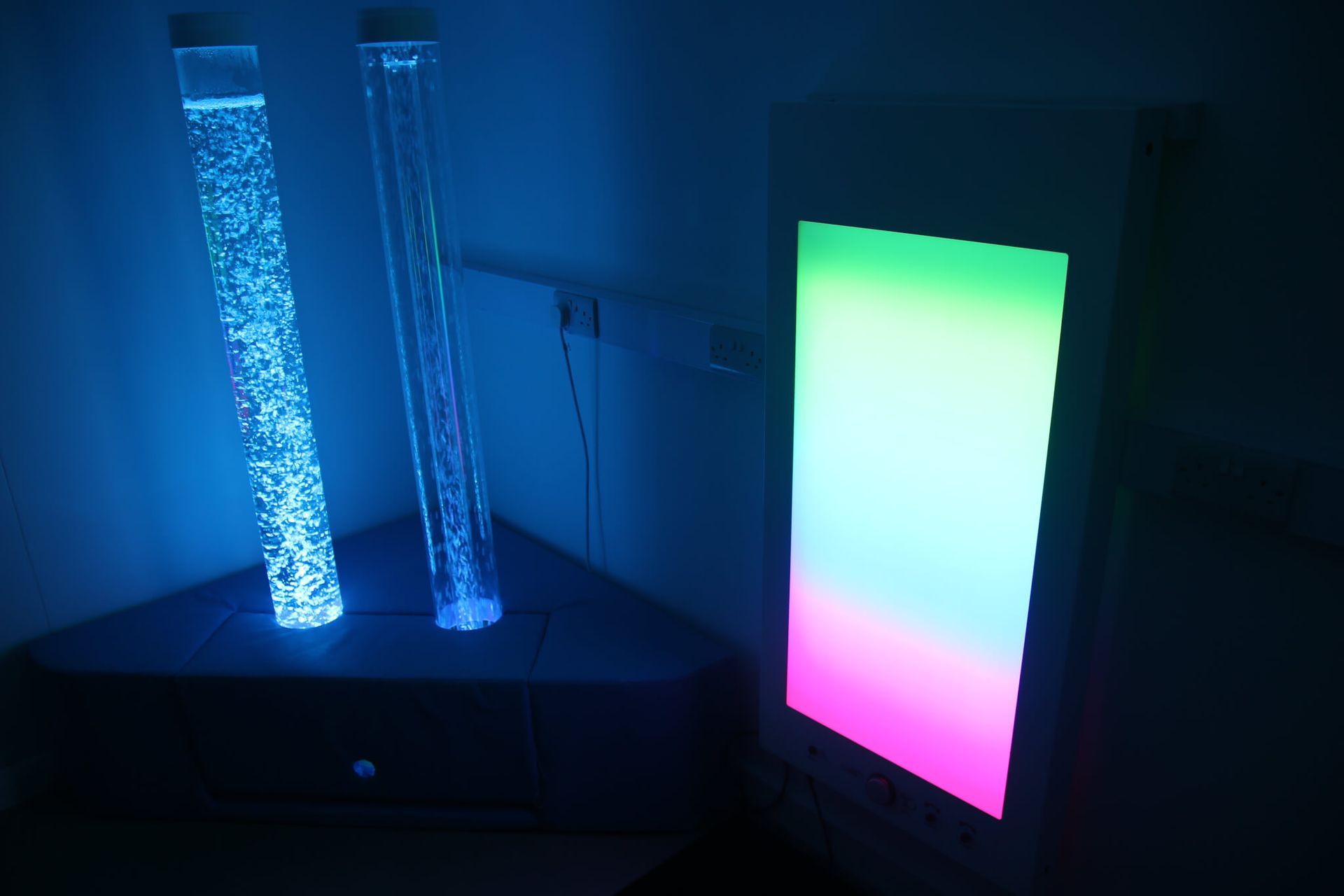 Two illuminated vertical bubble tubes and a glowing, multi-colored panel light in a dimly lit sensory room.