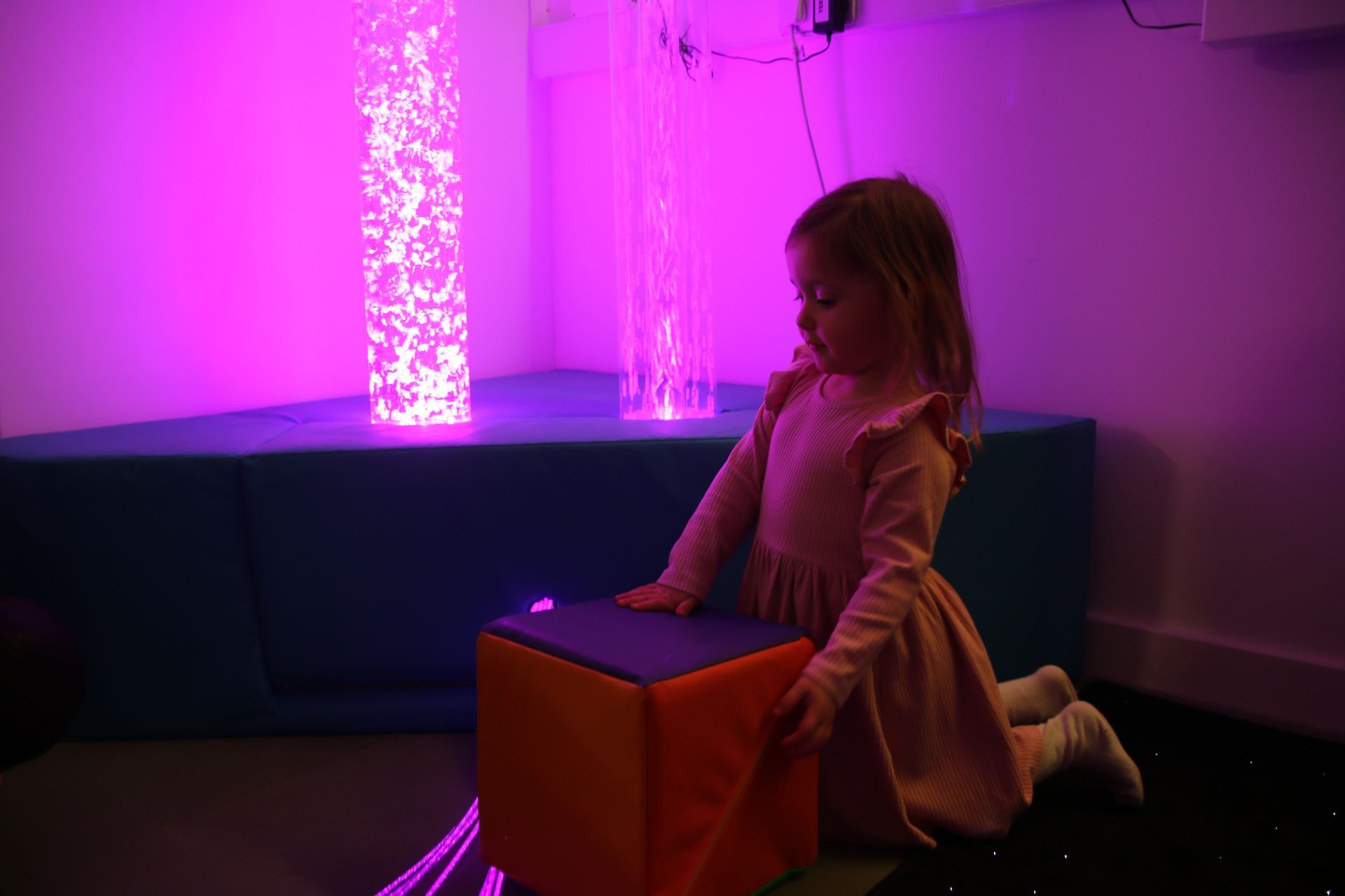 A child kneeling in a purple-lit sensory room, interacting with a colorful block in front of illuminated bubble columns.