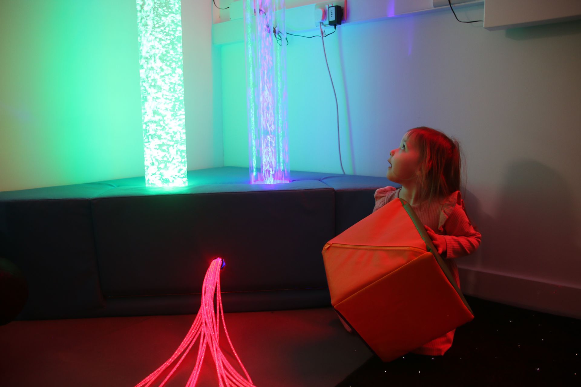 A child kneeling in a purple-lit sensory room, interacting with a colorful block in front of illuminated bubble columns.