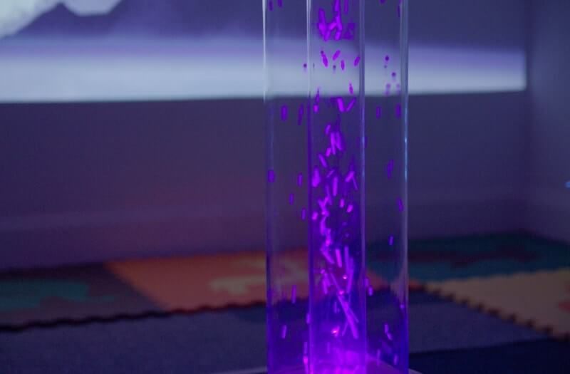 A purple bubble tube filled with rising bubbles, glowing against a blurred background in a sensory-friendly room.