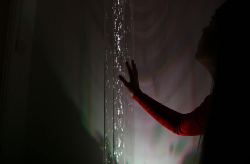 A purple bubble tube filled with rising bubbles, glowing against a blurred background in a sensory-friendly room.