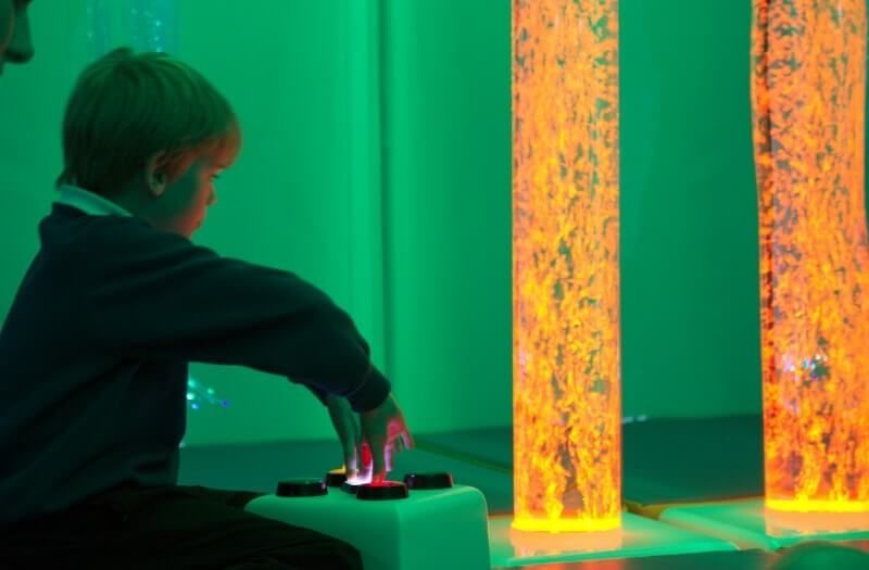 A person sitting in a green-lit sensory room, pressing a controller to interact with glowing, bubble-filled light columns.
