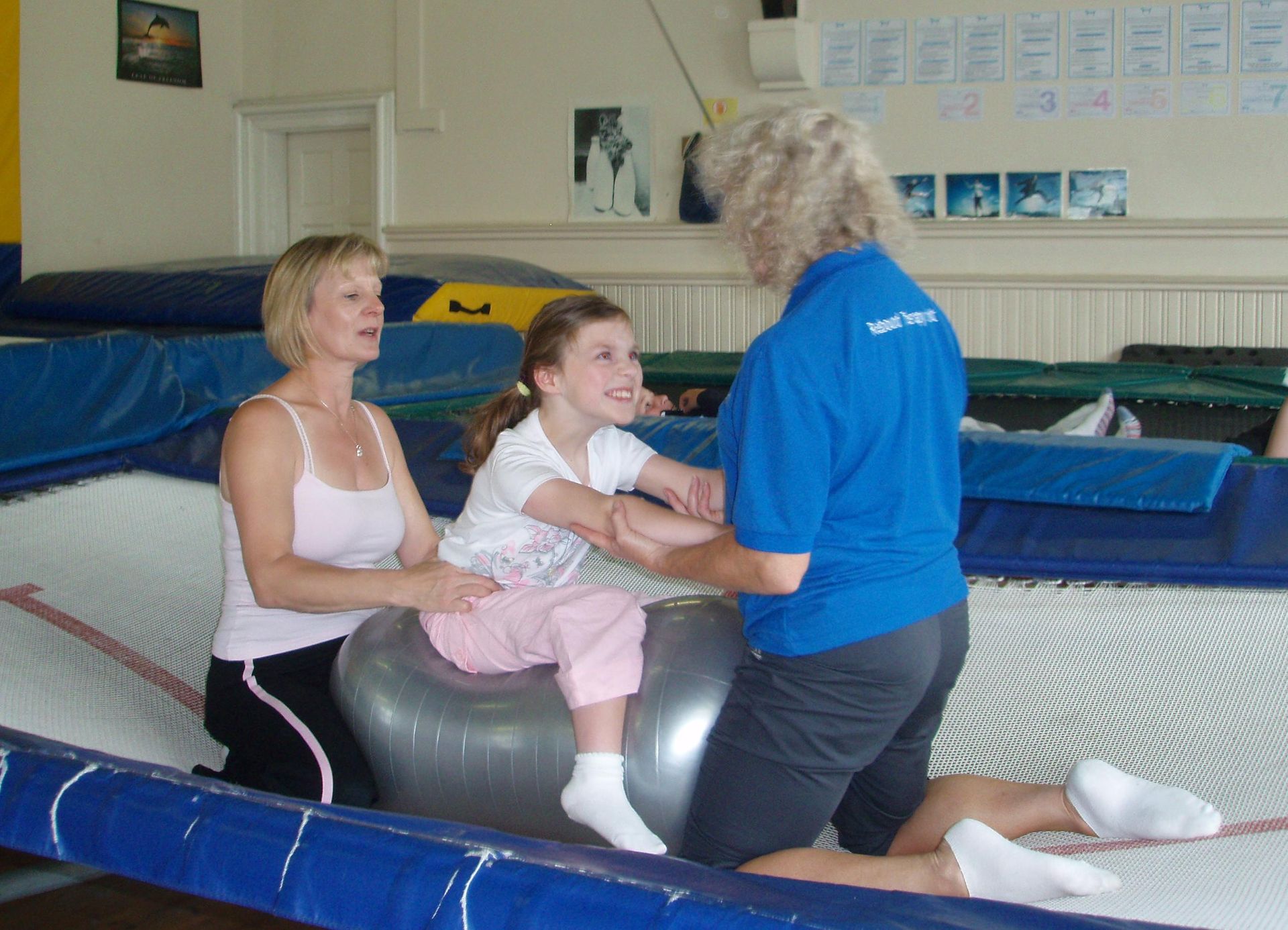 Two adults assist a child sitting on a grey exercise ball on a trampoline.