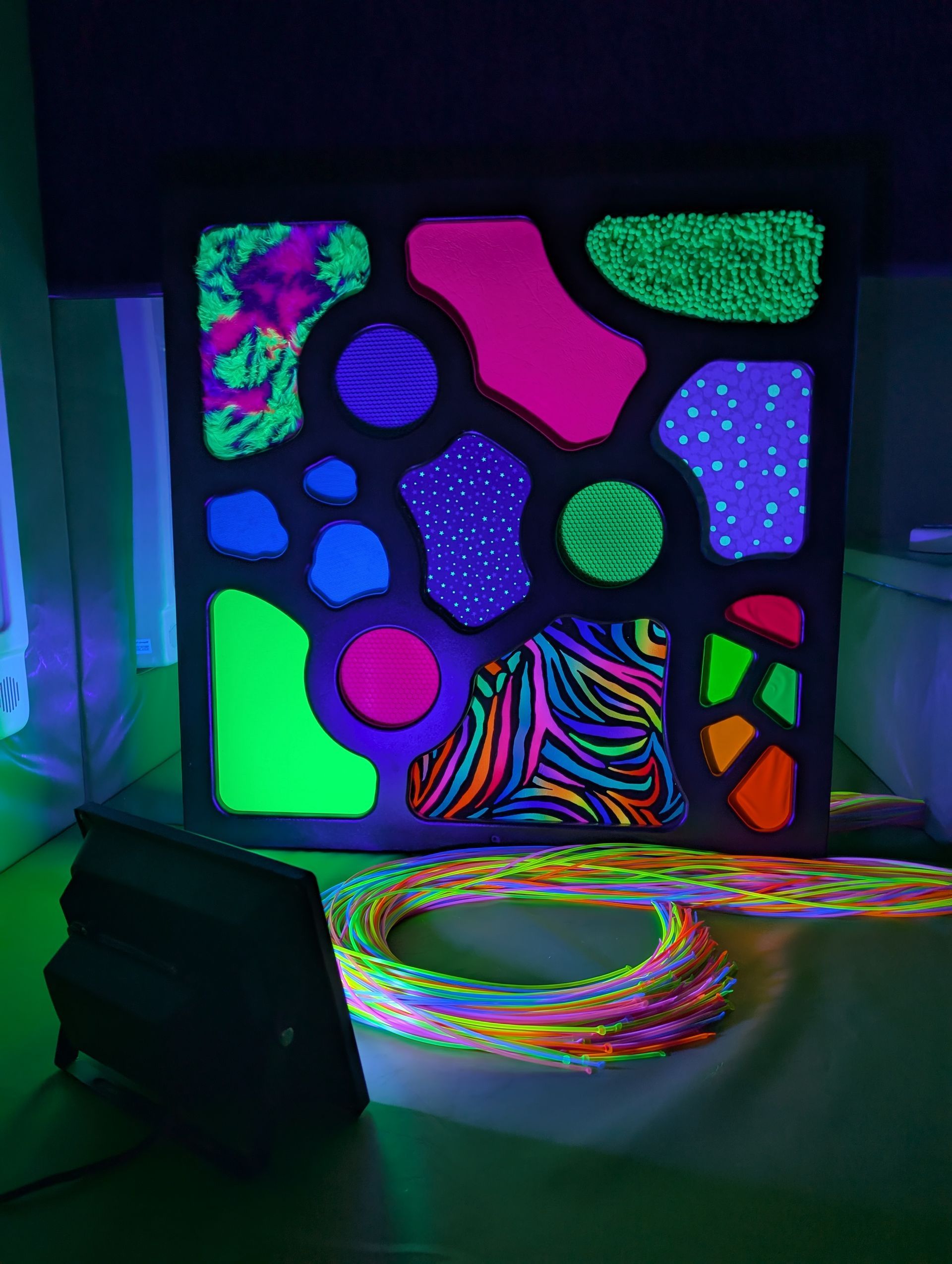 A black frame filled with vibrant, neon-colored patterns and shapes illuminated by a UV light, with coiled tubing below.