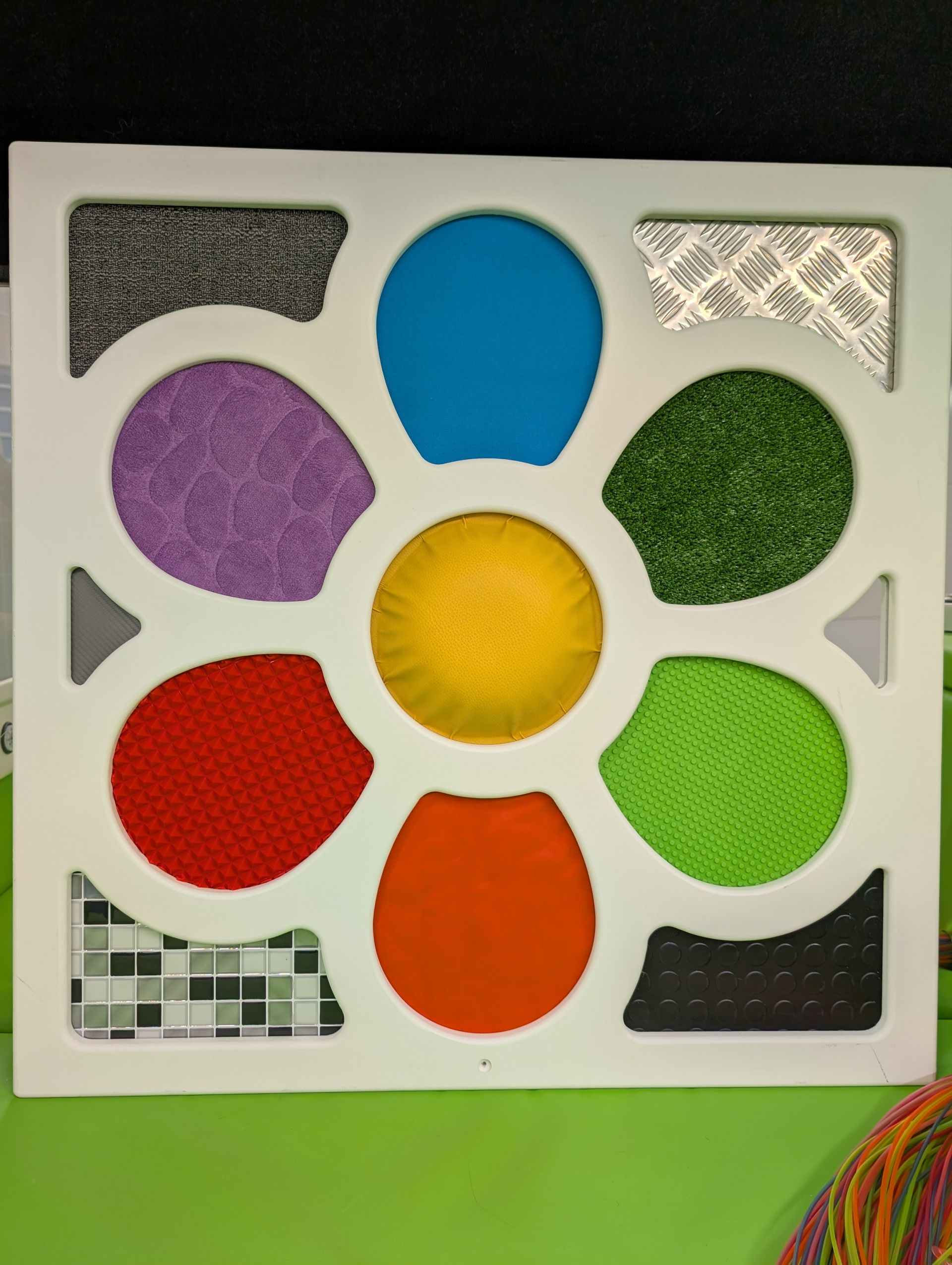 A square sensory board with a central yellow circle surrounded by six colorful, textured, petal-shaped fabric patches.