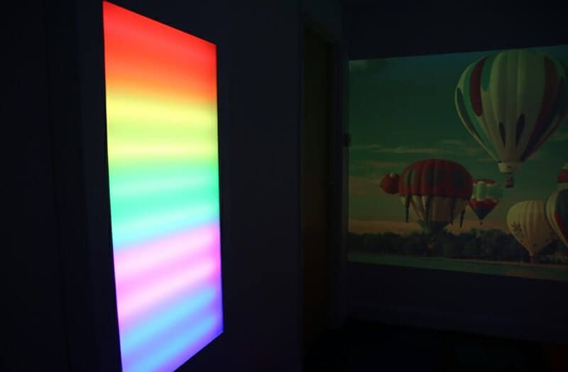 A vertical, glowing rainbow panel stands next to a projected scene of several hot air balloons in a dimly lit room.