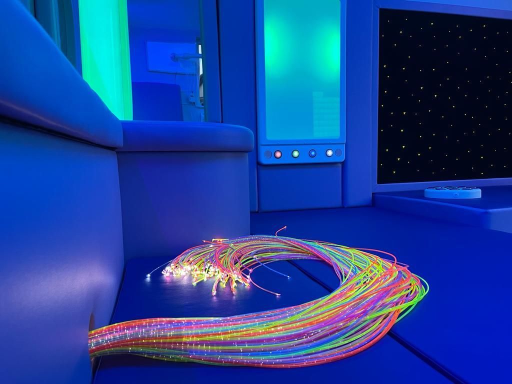 A cluster of glowing, multi-colored fiber optic strands rests on a blue surface in a sensory room with starry wall art.