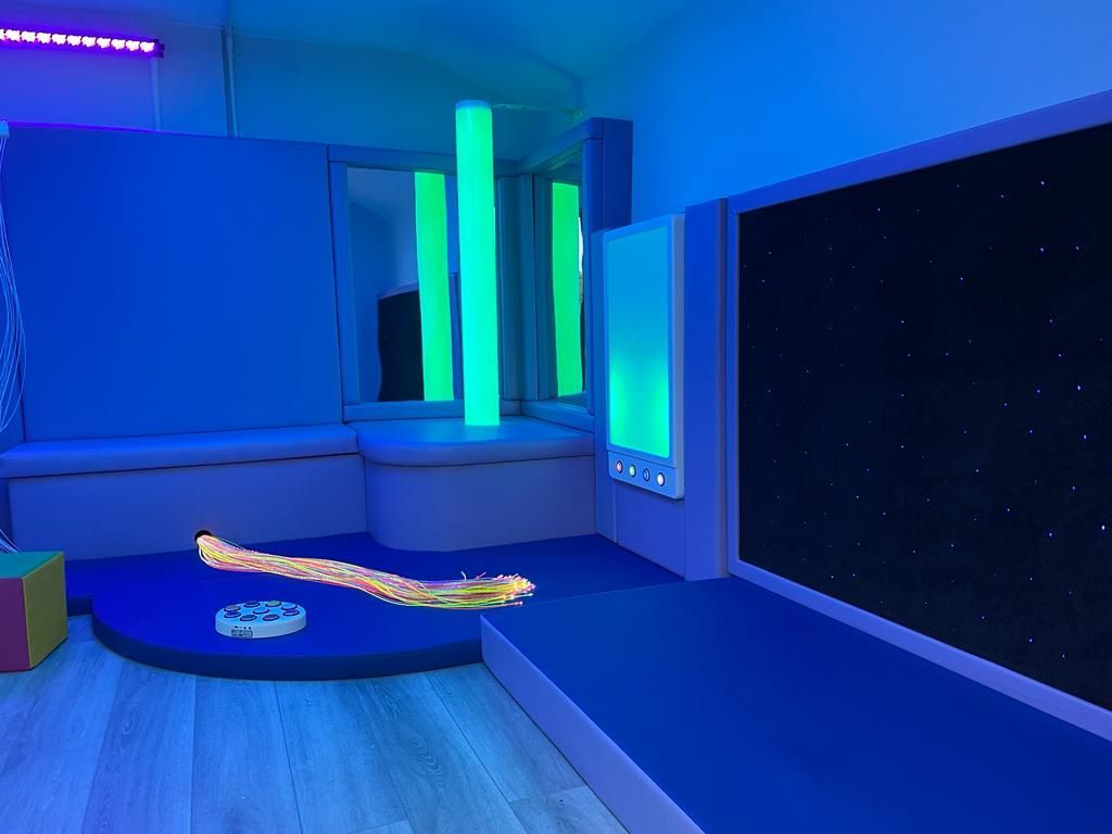 A sensory room bathed in blue light, featuring a glowing green bubble tube, a star-patterned wall, and soft floor mats.