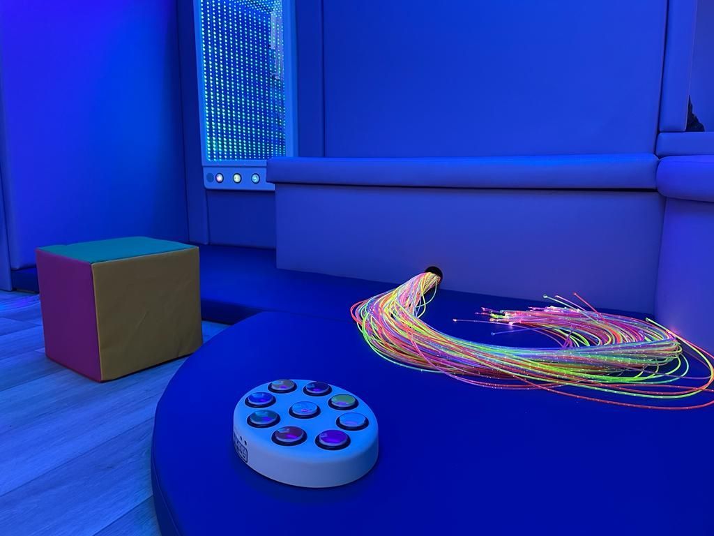 A sensory room with a colorful fiber optic light spray, a multi-button control pad, and a square block on a blue surface.