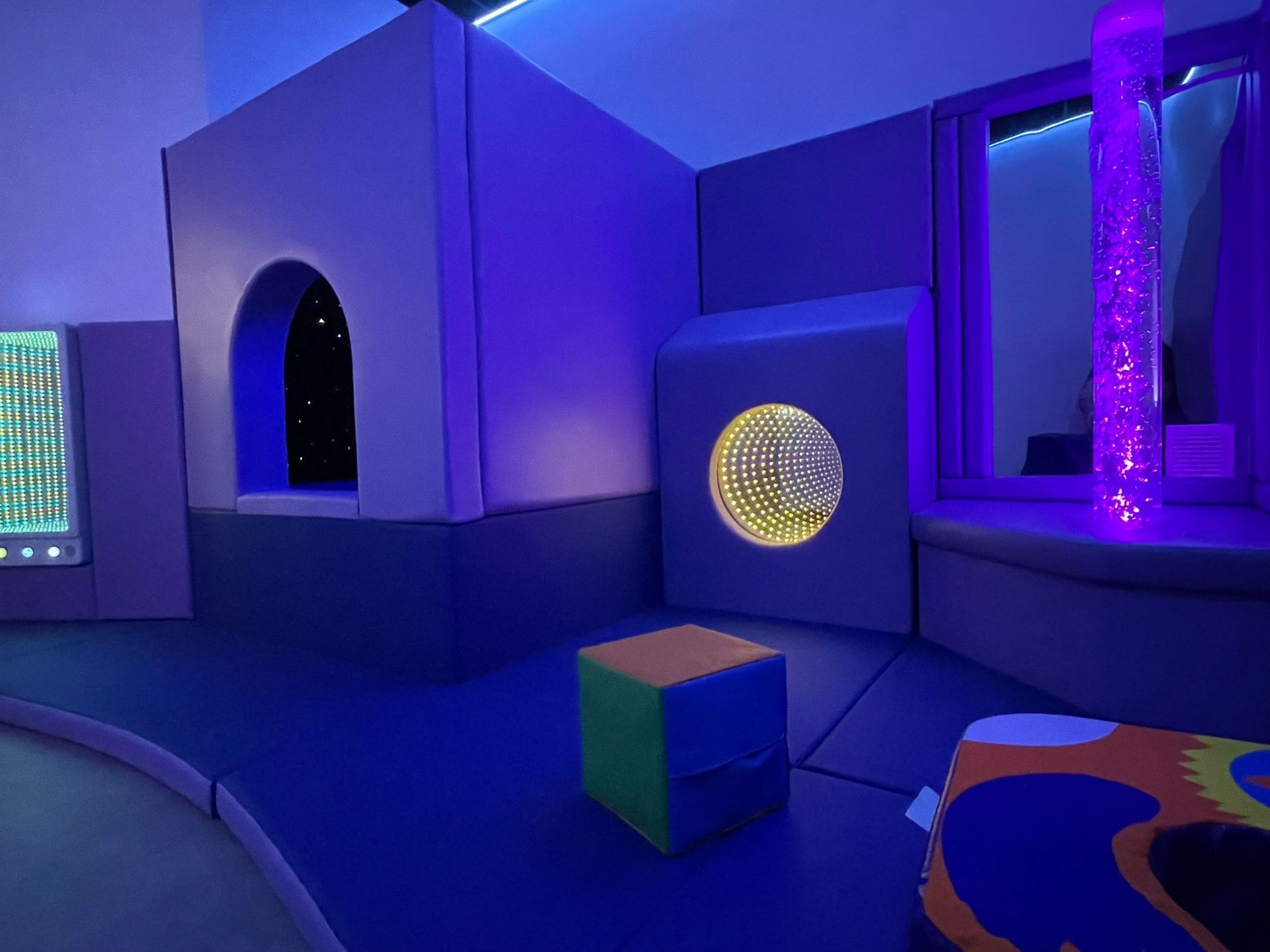 A sensory room with blue walls, padded structures, a light-up bubble tube, and a square green and orange foam block.