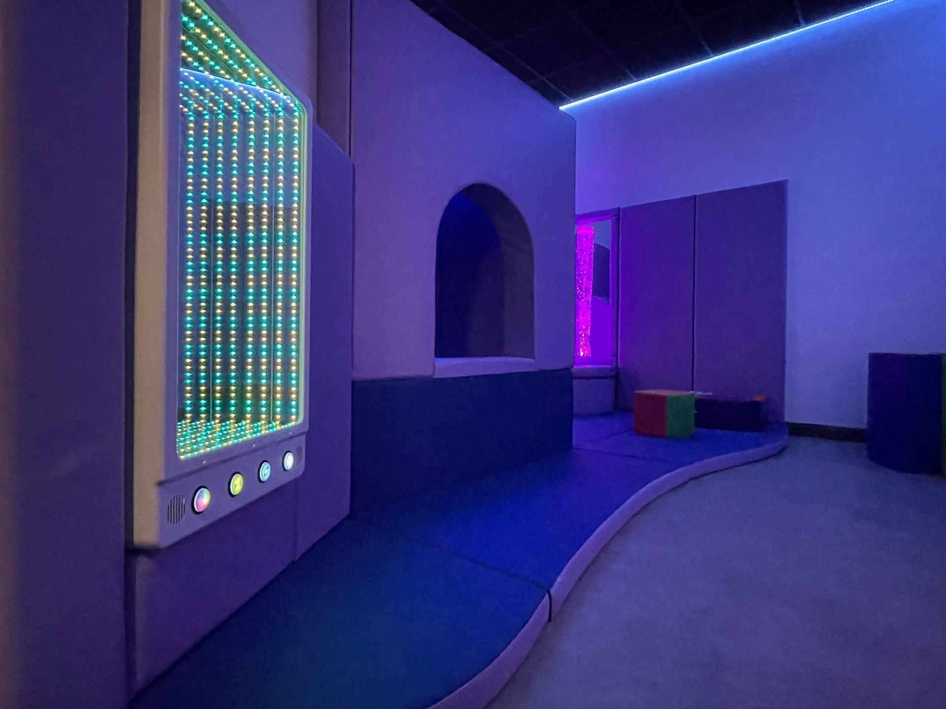 A sensory room with a glowing infinity mirror, blue padded flooring, an arched doorway, and purple accent lighting.
