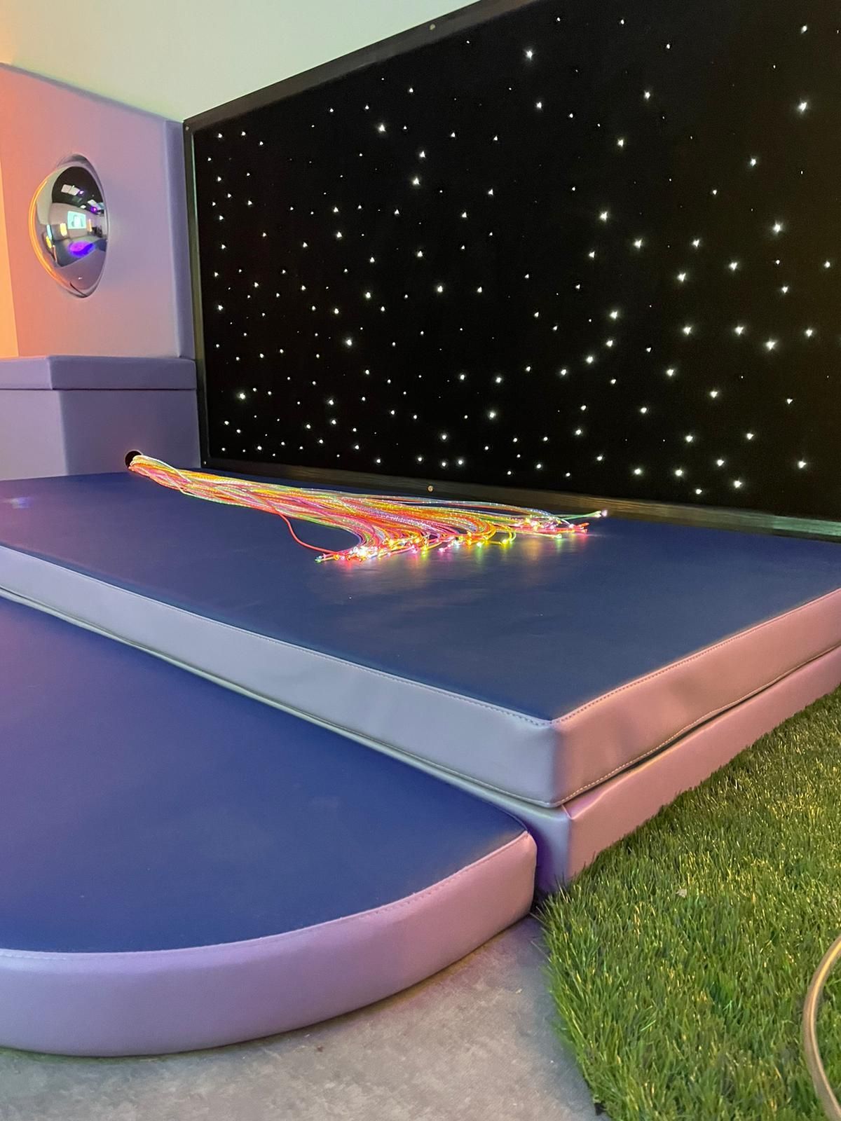 A multi-sensory room featuring a blue padded platform with fiber optic lights, a starry backdrop, and a mirror.