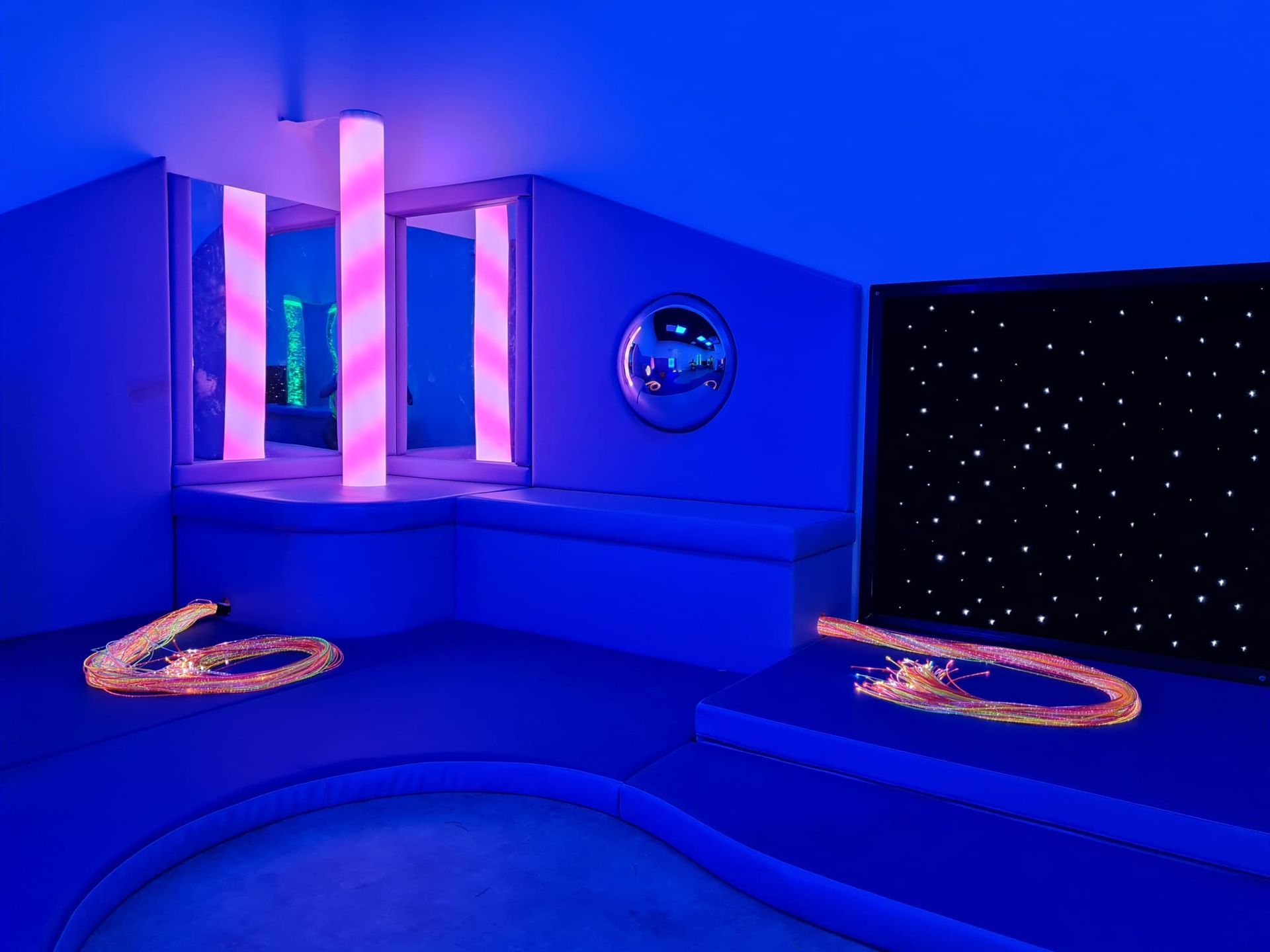 A sensory room with blue walls, a star-lit wall panel, pink-lit columns, and two fiber-optic light strands on platforms.