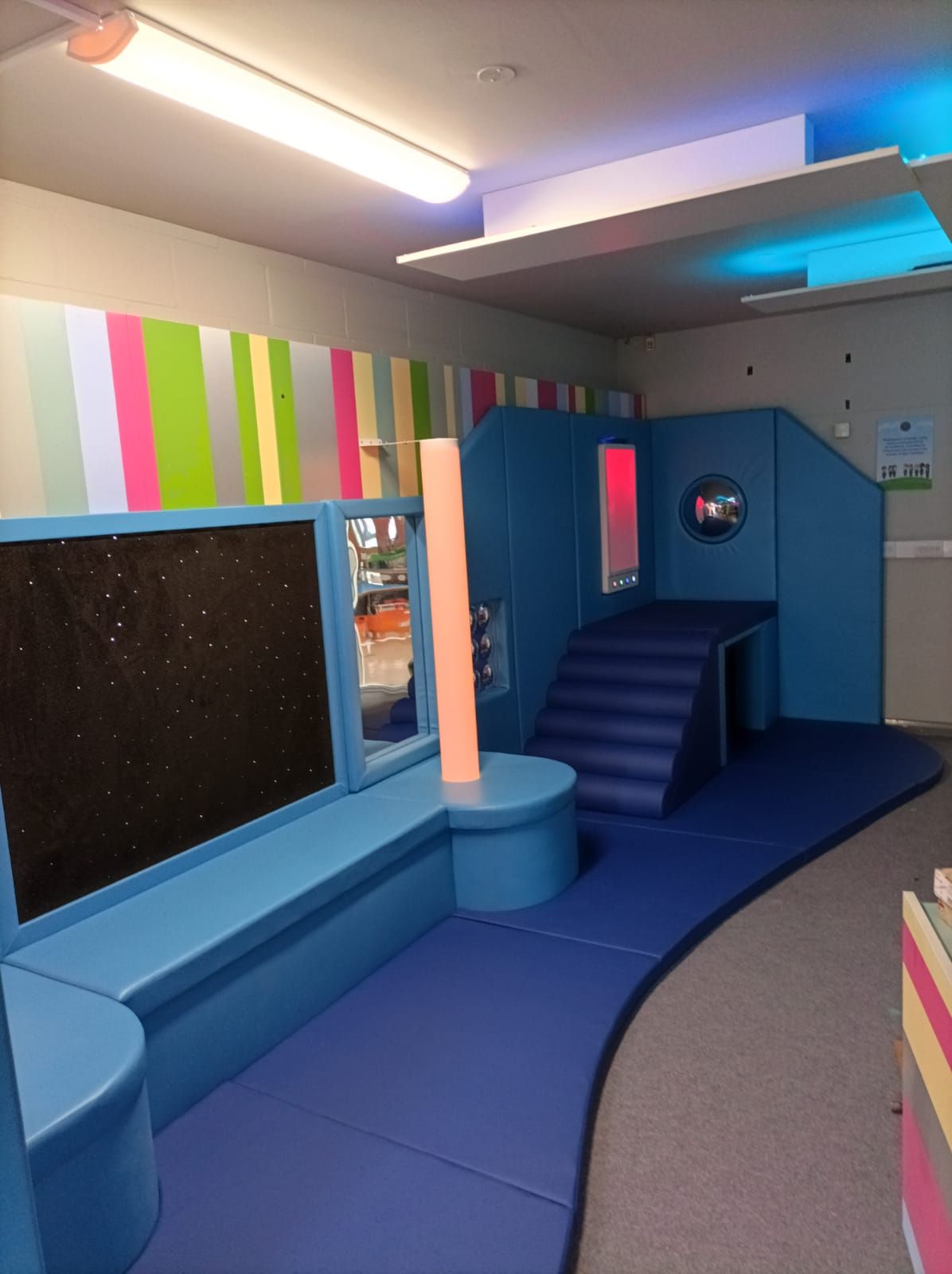 An indoor sensory play area with a blue padded structure, stairs, a dark wall panel, and multicolored striped wallpaper.