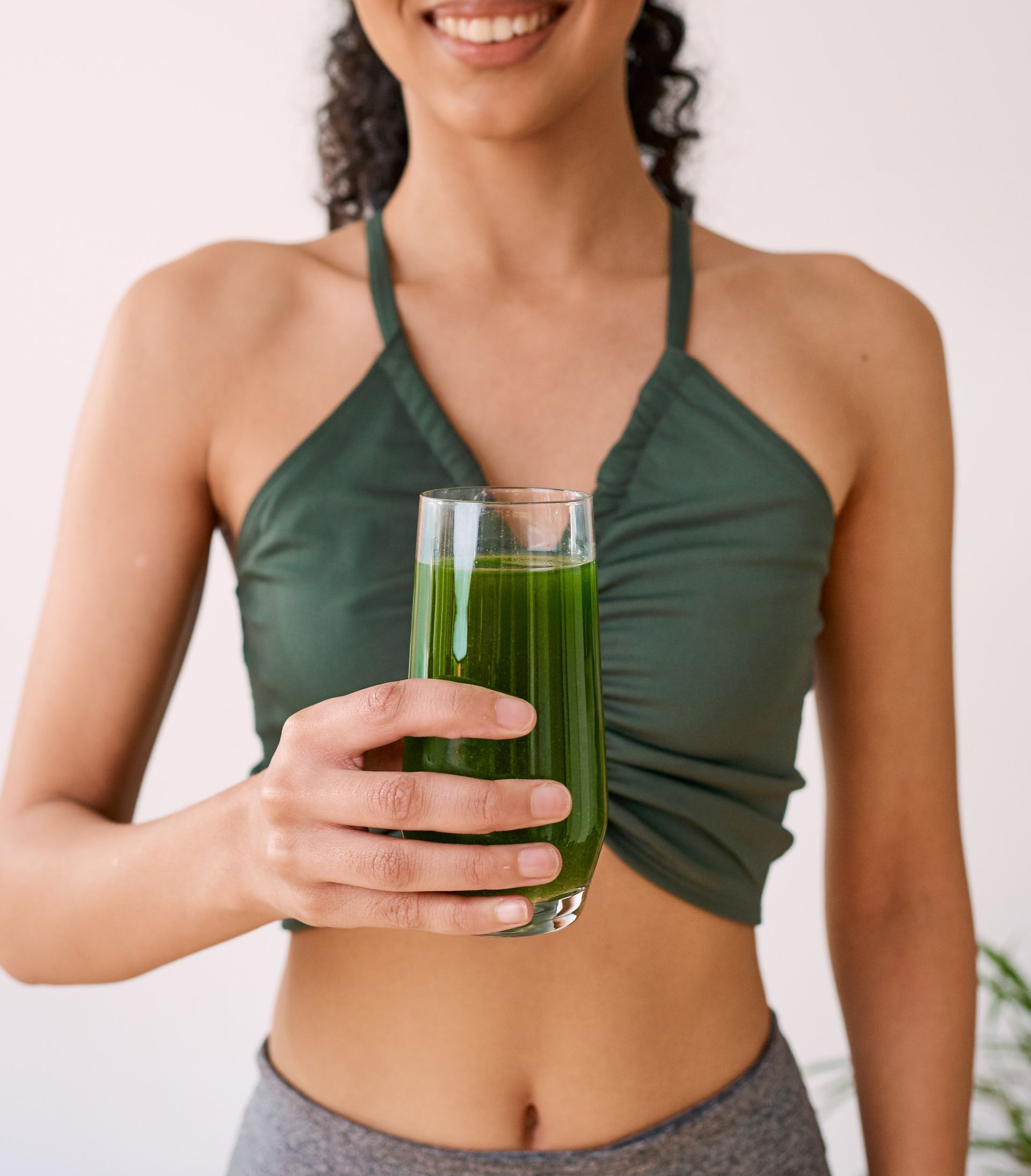 Woman holding a glass of green juice in a green halter top and gray leggings, cropped at the shoulders Woman holding a glass of green juice in a green halter top and gray leggings, cropped at the shoulders