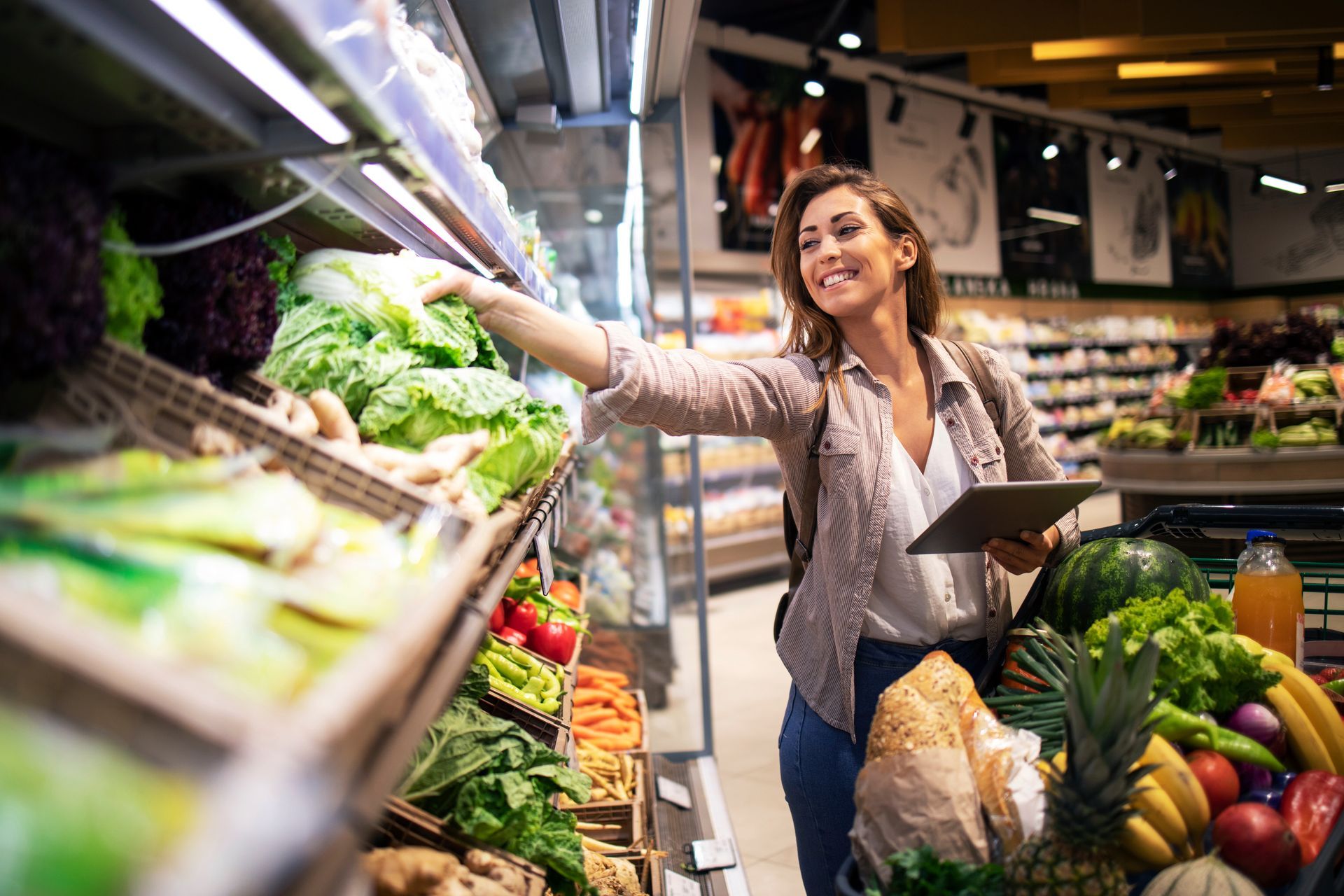 A store with a lot of food and a plant in the corner. Person selecting leafy greens in a grocery store while holding a tablet.