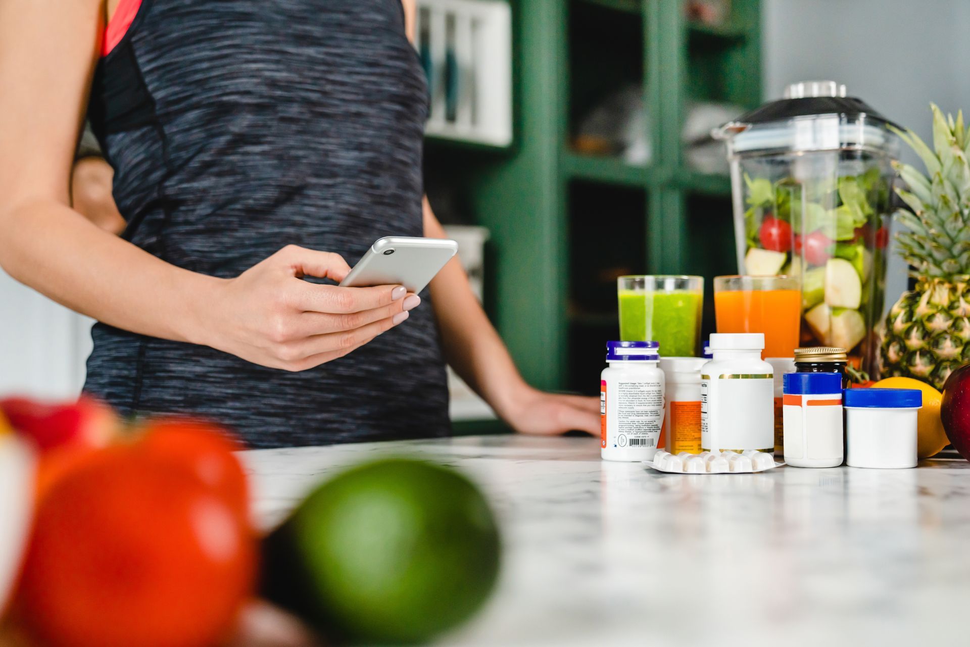 Person checking a smartphone beside a blender, produce, and supplement bottles.